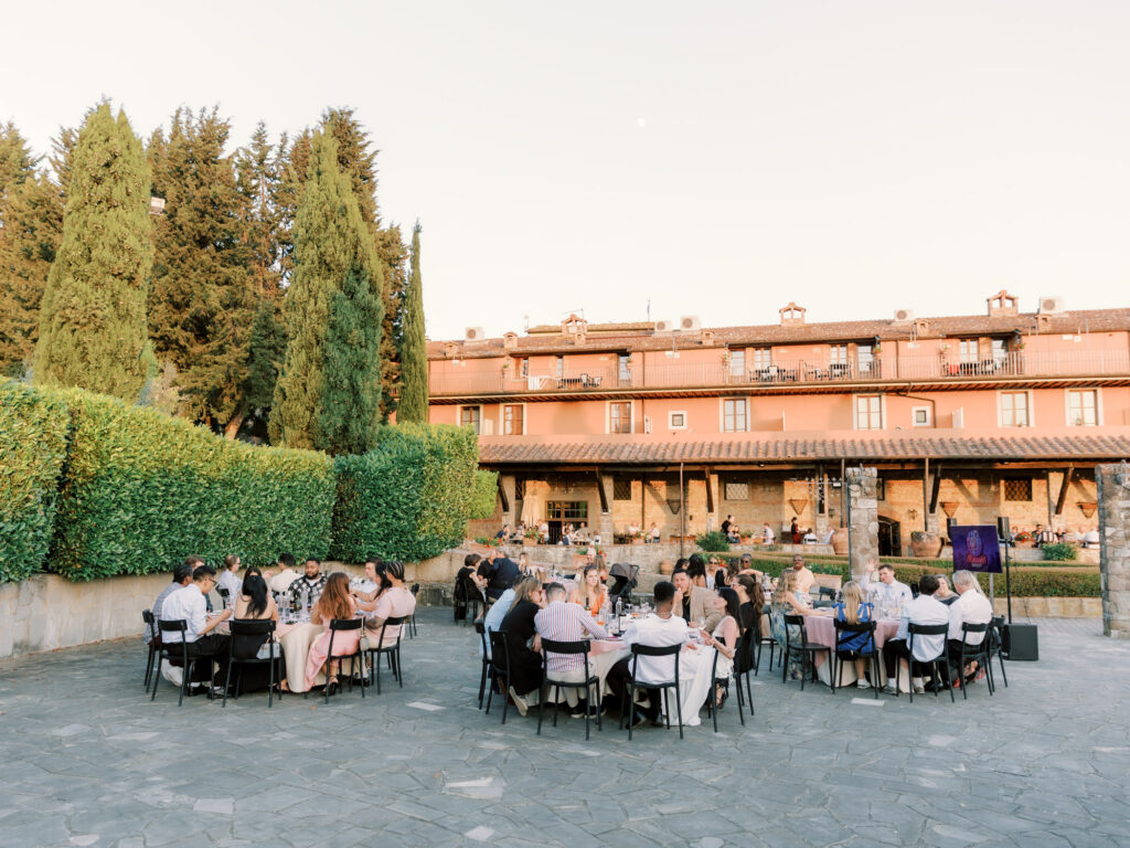 Wedding guests dressed in elegant semi-formal attire are seated at round tables with black chairs on a stone courtyard terrace at golden hour, with a glowing peach sky overhead. A tall manicured hedge and towering cypress trees frame the left side, while the warm terracotta facade of a rustic Tuscan villa with balconies and a covered loggia fills the background. A screen displaying a portrait is visible to the right of the dining area.