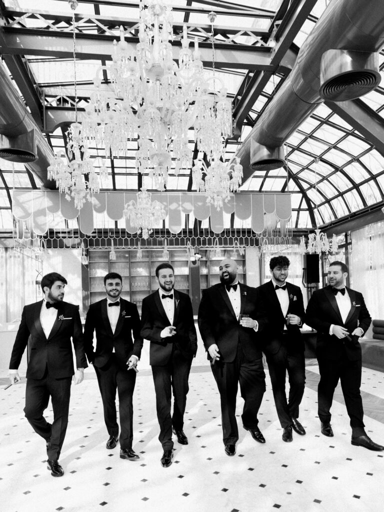 A striking black-and-white photograph of the groom and five groomsmen in matching black tuxedos and bow ties walking confidently side by side across a marble tiled floor inside the grand glass-roofed interior of The Address Montgomerie Dubai, with cascading crystal chandeliers hanging dramatically above them and an elegant bar backdrop behind them.