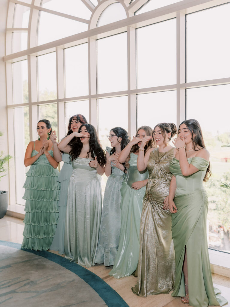 A candid emotional moment as six bridesmaids in mismatched sage green and gold floor-length gowns react with tears, laughter, and joy upon seeing the bride for the first time, gathered together in front of the floor-to-ceiling windows of The Address Montgomerie Dubai with the golf course greenery softly glowing behind them.