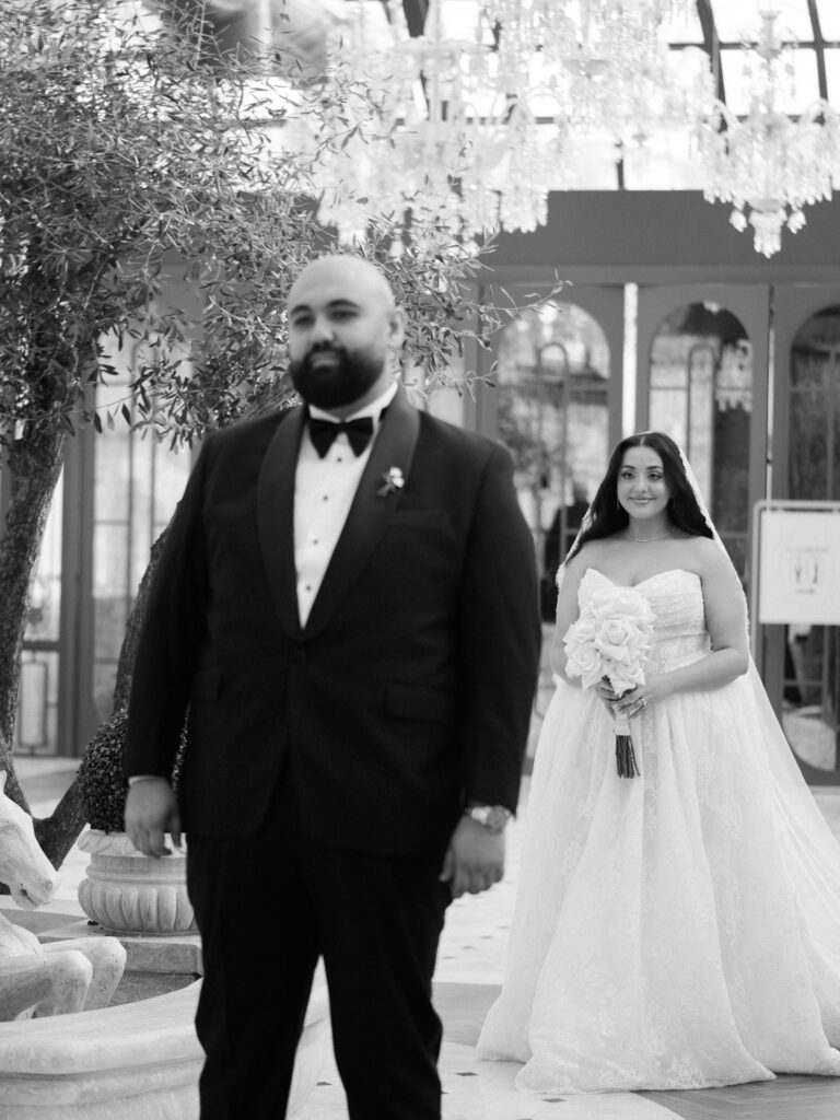 A black-and-white first look moment as the groom in his black tuxedo turns to see the bride in her voluminous white off-the-shoulder ballgown and cathedral veil for the first time inside the grand glass-roofed interior of The Address Montgomerie Dubai, his expression overcome with emotion.

