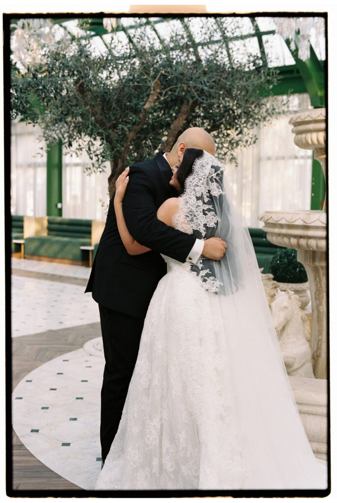 The groom embraces the bride from behind in an emotional first hug after their vows inside the glass-roofed atrium of The Address Montgomerie Dubai, the bride's lace cathedral veil and voluminous ballgown train spreading across the marble floor, with olive trees and crystal chandeliers glowing above them.

