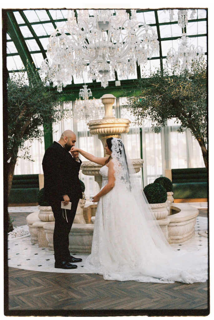 The groom gently places the veil over the bride's face in a tender ceremonial moment inside the grand glass-roofed atrium of The Address Montgomerie Dubai, with cascading crystal chandeliers, a stone fountain, and olive trees creating an opulent backdrop around them.

