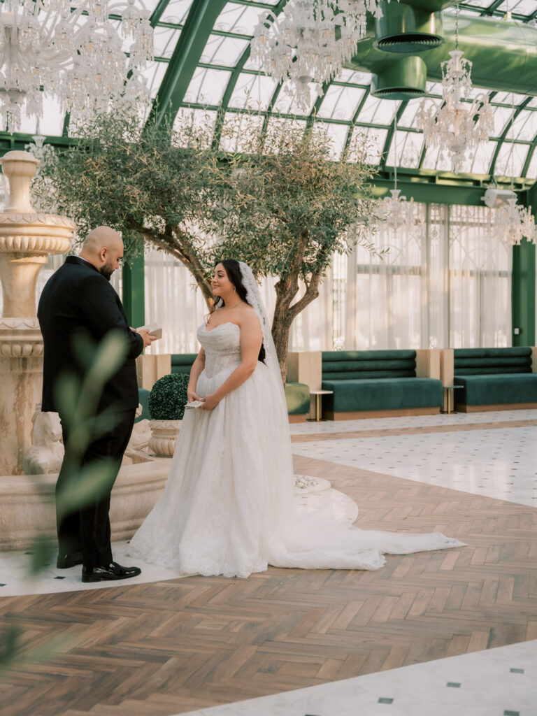 The bride and groom stand facing each other during their private vow exchange inside the elegant glass greenhouse interior of The Address Montgomerie Dubai, surrounded by olive trees, green panelled walls, and warm parquet flooring, the bride's cathedral veil and ballgown train spreading across the floor.

