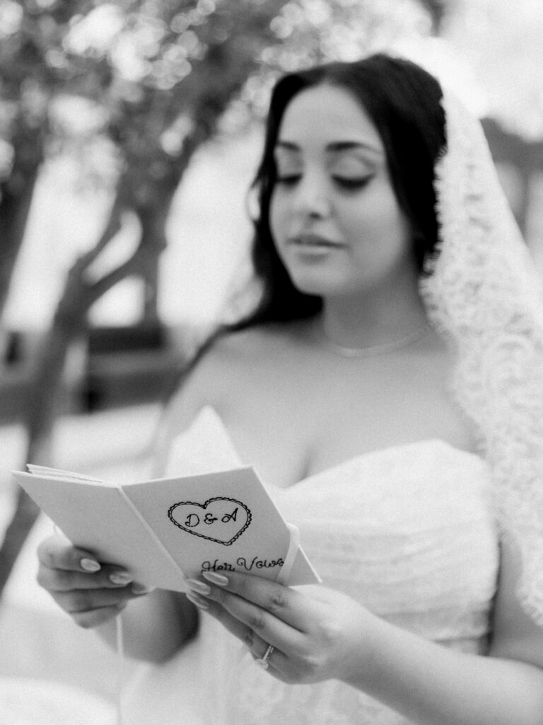 A black-and-white close-up of the bride in her off-the-shoulder lace ballgown reading her handwritten wedding vows from a small booklet, her expression focused and emotional during the private ceremony at The Address Montgomerie Dubai.

