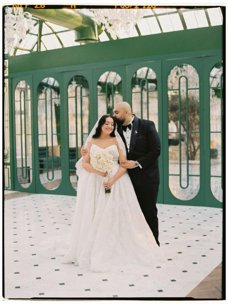 A film photograph of the bride and groom standing close together and smiling inside The Address Montgomerie Dubai's elegant green-panelled glass corridor, the bride in her white ballgown holding her white bouquet and the groom in his black tuxedo, with the geometric marble floor stretching before them.