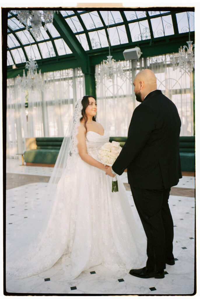 The bride stands reading her vows while the groom listens attentively inside the glass-roofed atrium of The Address Montgomerie Dubai, crystal chandeliers and olive trees visible above them, the bride's long cathedral veil and ballgown train pooling gracefully across the marble floor.
