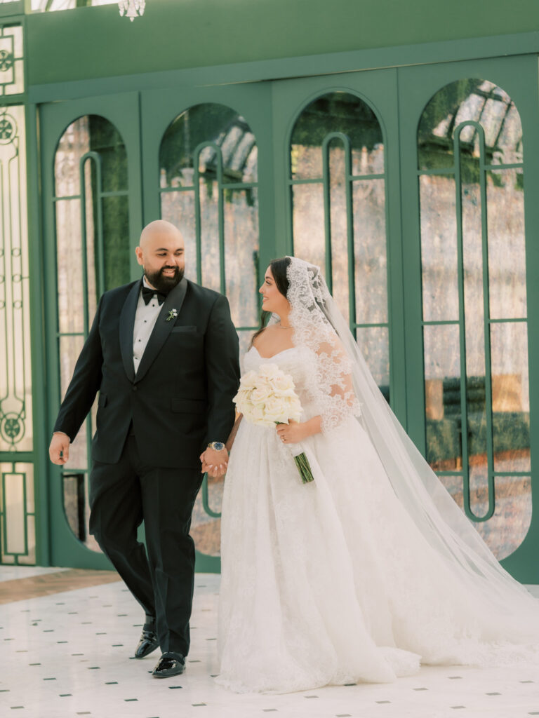 The bride and groom walk hand in hand laughing through the grand green-arched glass corridor of The Address Montgomerie Dubai, the bride's voluminous white ballgown and cathedral veil flowing elegantly behind her as the groom smiles adoringly at her.