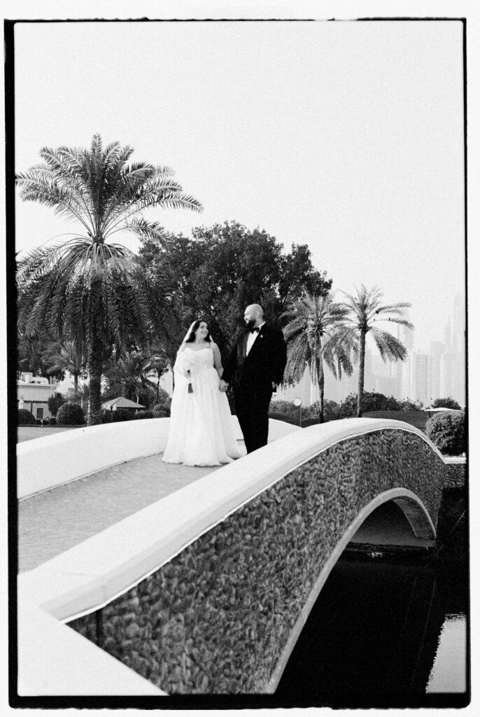 A black-and-white wide shot of the bride and groom standing together on a curved stone bridge at The Address Montgomerie Dubai, framed dramatically by towering palm trees, the bride's voluminous ballgown and veil cascading down the bridge steps in elegant contrast to the soft sky above.