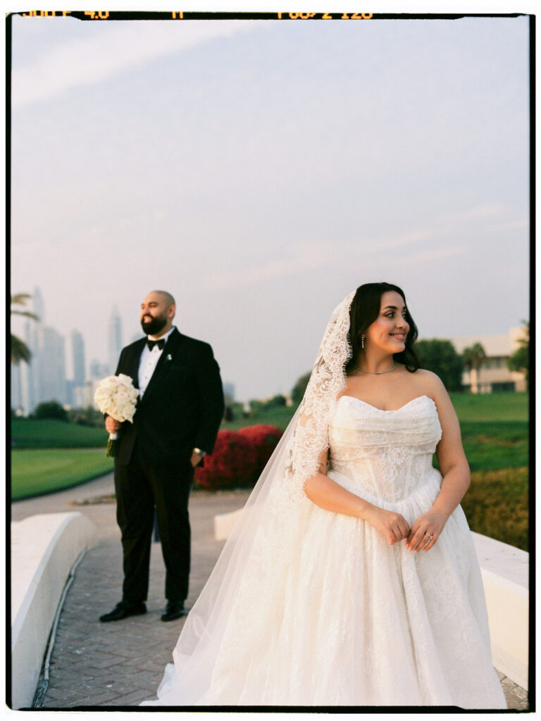 The bride and groom stand together on the golf course grounds of The Address Montgomerie Dubai at dusk, both smiling warmly at the camera, with the iconic Dubai skyline — including recognisable high-rise towers — glowing softly in the dusky blue background behind them and red floral hedges adding colour to the scene.
