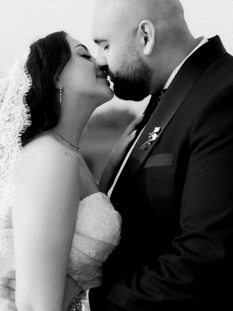 A close-up black-and-white photograph of the bride and groom sharing a passionate kiss, the bride in her lace-detailed off-the-shoulder ballgown and the groom in his black tuxedo, captured in a deeply intimate and cinematic moment at The Address Montgomerie Dubai wedding.