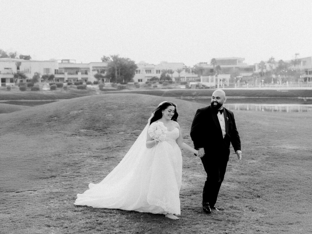A black-and-white photograph of the bride and groom walking hand in hand across the manicured fairways of The Address Montgomerie golf course in Dubai, both laughing joyfully. The bride wears a voluminous off-the-shoulder ballgown and holds a rounded bouquet, her long train sweeping across the grass, while the groom in a black tuxedo and bow tie smiles adoringly at her, with the golf course water feature and Dubai residential villas softly visible in the background.