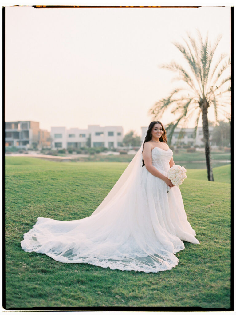 

The bride stands alone on the lush green fairways of The Address Montgomerie Dubai golf course at golden hour, her voluminous white off-the-shoulder ballgown and long cathedral veil spreading dramatically across the grass, holding her white bouquet with palm trees and modern Dubai villas softly visible behind her.