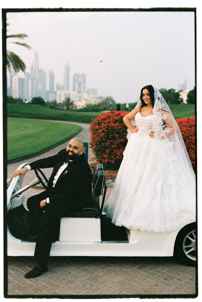 The bride and groom pose confidently beside a luxury white car at The Address Montgomerie Dubai, the groom leaning against the door in his black tuxedo while the bride stands beside him in her voluminous off-the-shoulder ballgown and lace cathedral veil, with red floral hedges and the Dubai skyline glowing in the background.