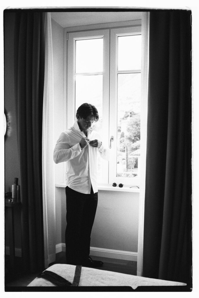 A black-and-white photograph of the groom standing in profile at a bright window, buttoning his white dress shirt as he gets ready on his wedding day, with the lush Lake Como hillside softly visible through the glass behind him.