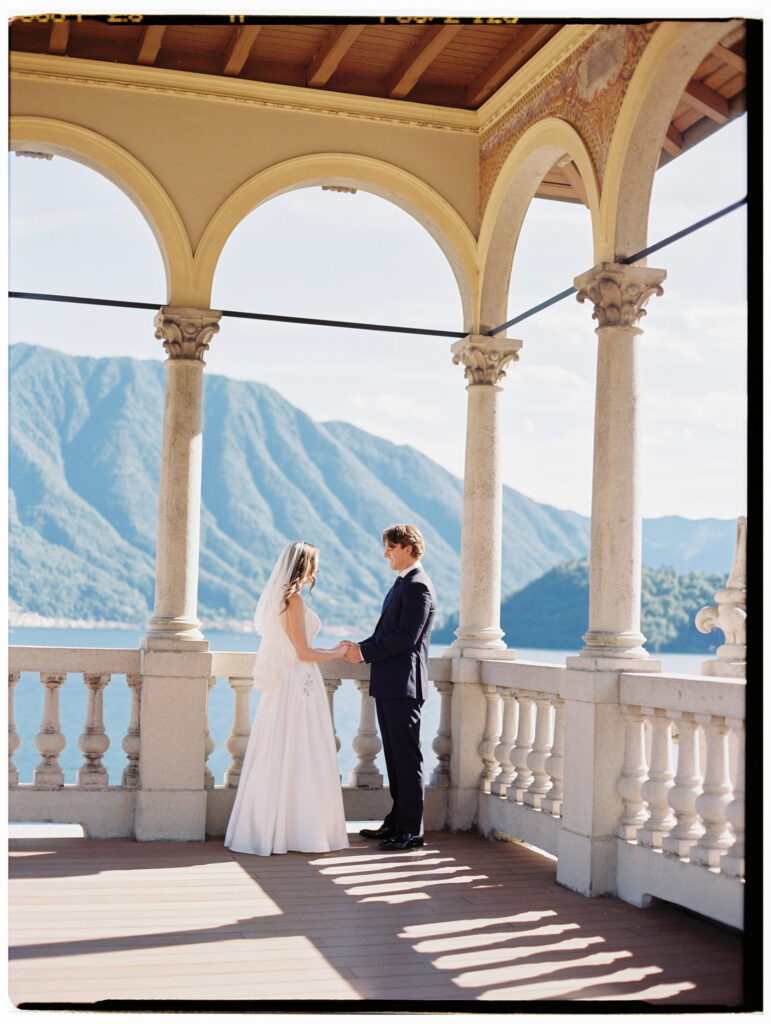 A wide shot of the bride and groom standing together on the ornate arched colonnade terrace of a Lake Como villa, dramatic stone pillars framing them against a breathtaking view of the Alpine mountains and shimmering blue lake.