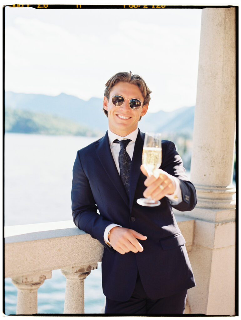 The groom in a navy suit and sunglasses raises a champagne flute toward the camera with a confident, celebratory smile, leaning against the stone balustrade of the Lake Como villa terrace with the Alpine mountains and blue lake filling the bright background behind him.