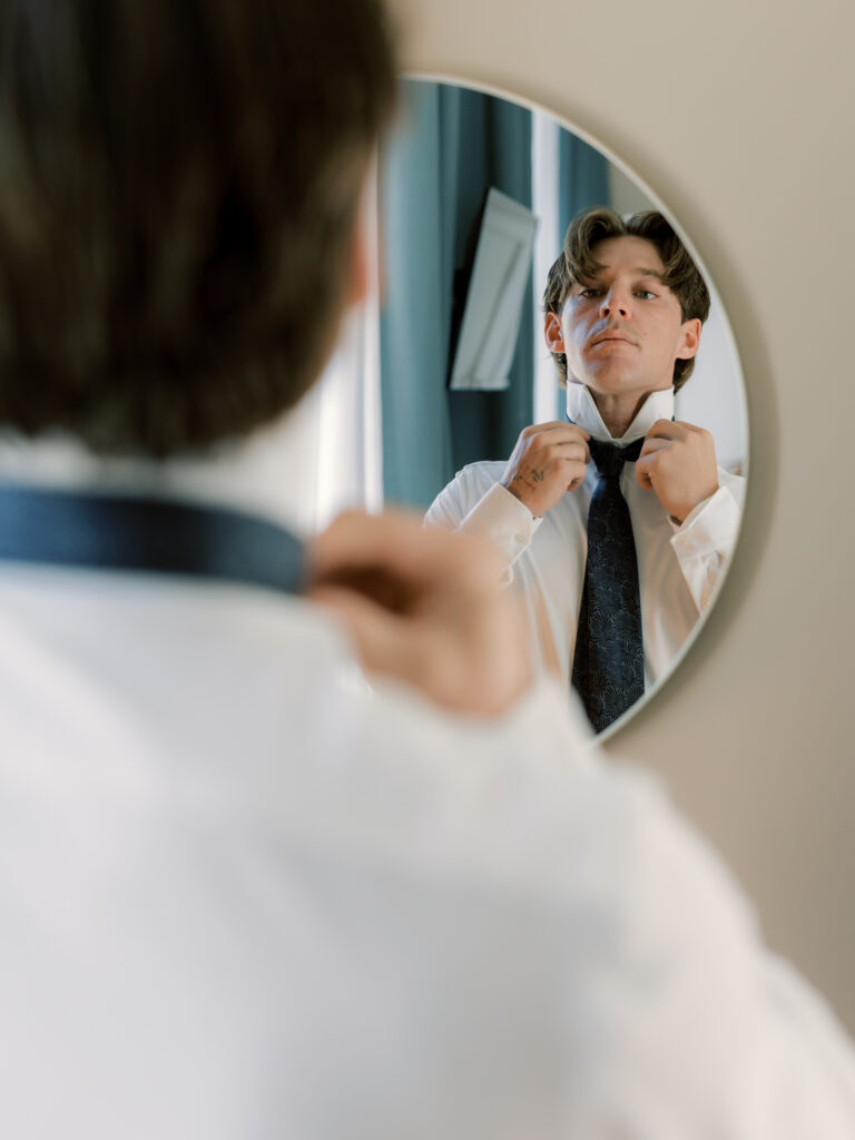 A close-up photograph of the groom adjusting his dark tie in a round mirror, his expression focused and composed as he finishes getting ready inside their Lake Como hotel room.