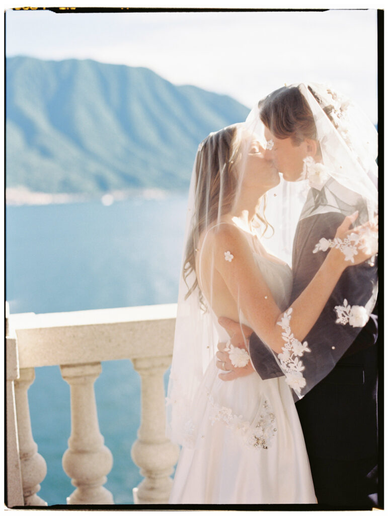 A tender close-up film photograph of the groom in his navy suit embracing the bride from behind on the Lake Como villa terrace, her floral-embroidered cathedral veil draped over his arm as they share a quiet, intimate moment with the blue lake shimmering behind them.