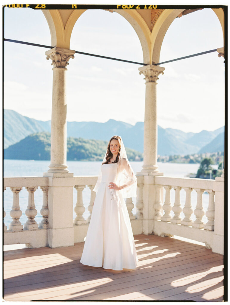 A wide architectural shot of the bride standing alone at the far end of the grand arched colonnade terrace of the Lake Como villa, her white gown a small, elegant figure framed by the repeated stone arches and the vast mountain and lake panorama beyond.