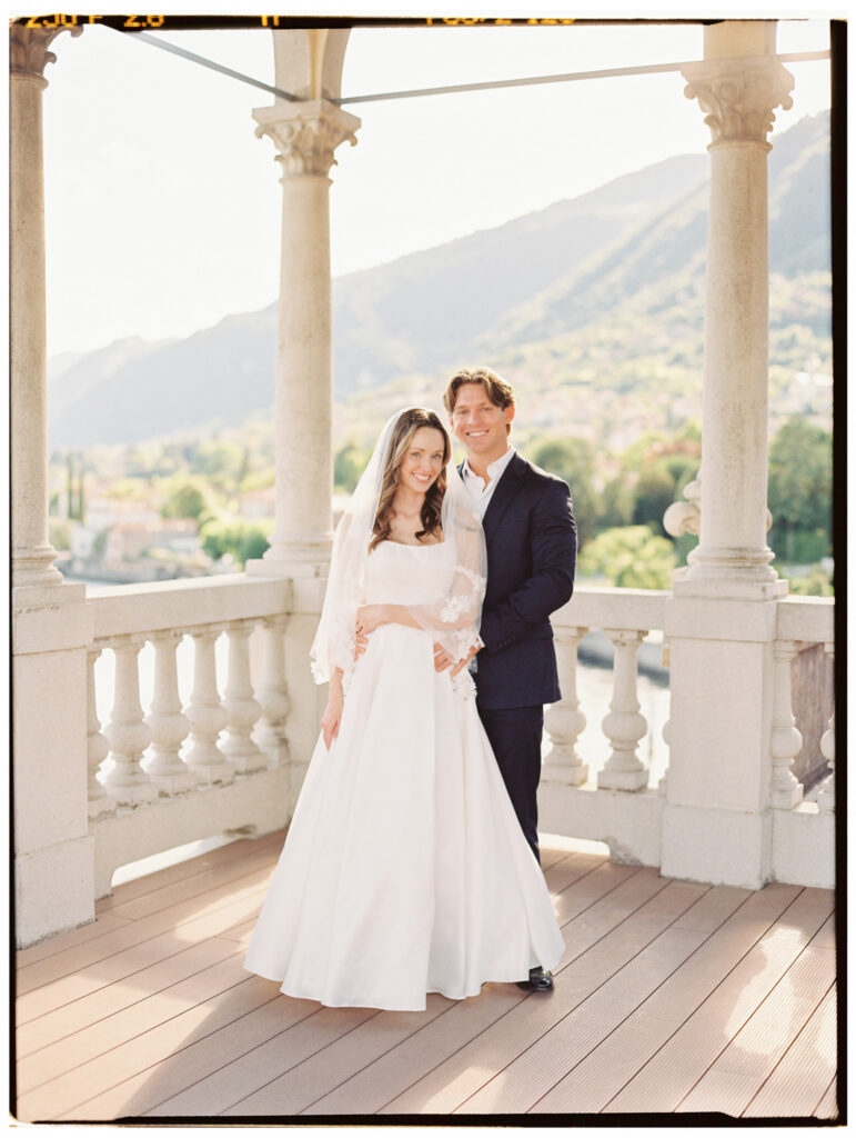 The bride and groom stand together smiling warmly on the sunlit wooden deck of the Lake Como villa terrace, the bride in her long-sleeved lace overlay wedding gown and the groom in his navy suit, with the lake and mountain panorama glowing behind them.