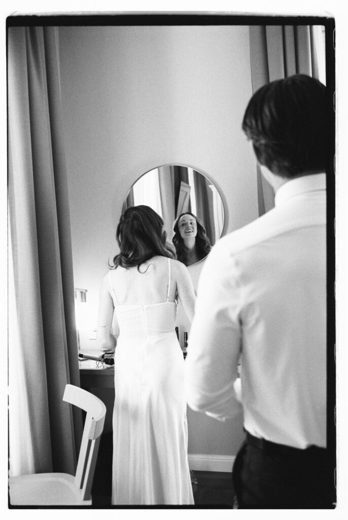 A black-and-white film photograph of the groom fastening the back of the bride's wedding dress as she looks at her reflection in a round mirror, captured in a soft, intimate getting-ready moment inside their Lake Como hotel room.