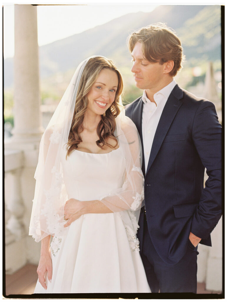 The bride and groom stand side by side on the sunlit Lake Como villa terrace, both smiling broadly at the camera, the bride in her long-sleeved lace wedding gown and the groom in his navy suit, with the lake and Alpine mountain vista glowing warmly behind them.