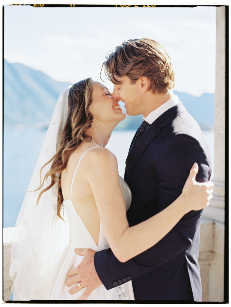 The bride and groom share a close, joyful moment — foreheads nearly touching and both smiling — on the sunlit stone terrace of a Lake Como villa, the bride in her white spaghetti-strap gown and cathedral veil, the groom in a navy suit.