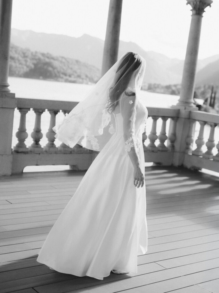 A black-and-white film photograph of the bride walking gracefully along the stone balustrade terrace of the Lake Como villa, her full white gown and veil trailing behind her in the sunlight, the arched columns casting dramatic shadows across the wooden deck.