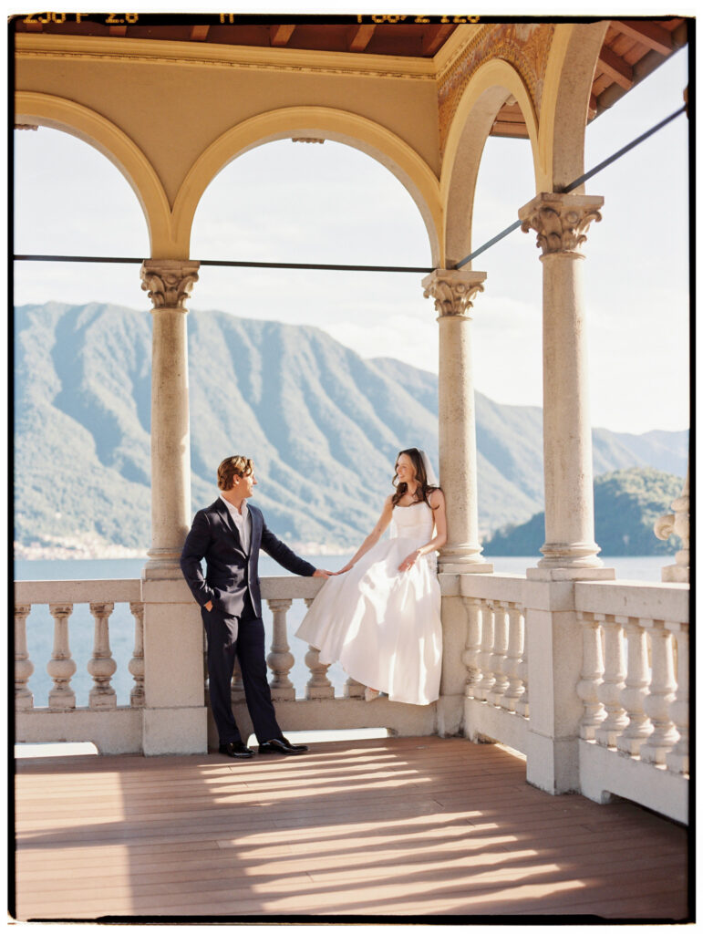 The groom leads the bride by the hand across the ornate arched colonnade terrace of the Lake Como villa, both in profile against the stunning backdrop of the Alpine mountains and glittering lake waters visible through the stone arches.

