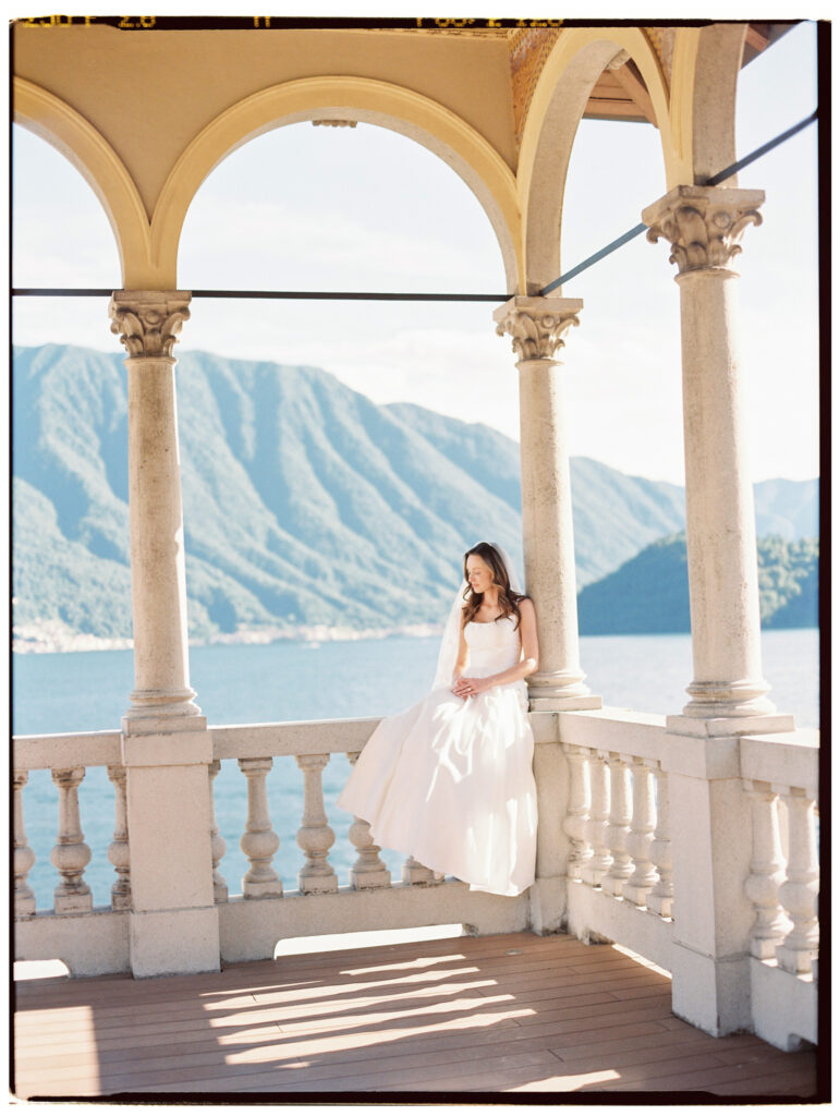 The bride sitting on the balustrade on a  arched colonnade terrace of a Lake Como villa, her flowing white gown and veil catching the breeze, with the vivid blue lake and steep green Alpine mountains stretching dramatically behind her through the stone arches.
