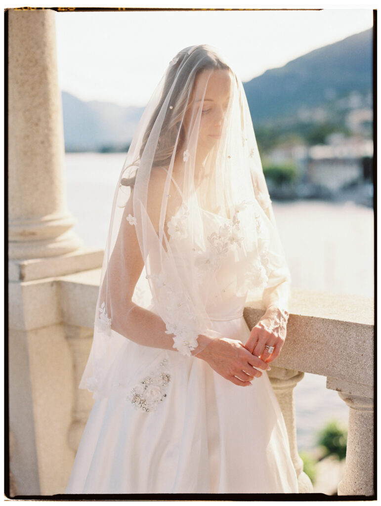 A close-up detail shot of the bride's white wedding gown, showcasing delicate floral embroidery along the skirt hem and her cathedral veil cascading softly beside it in bright natural light.