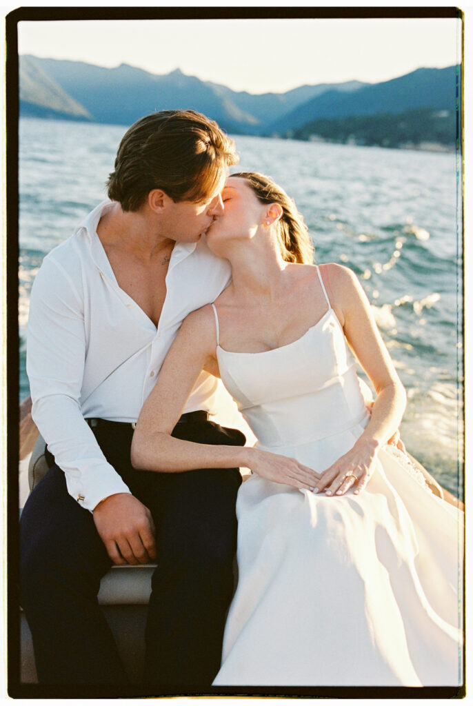 The bride and groom share a gentle kiss aboard the classic wooden Riva speedboat on Lake Como, the bride in her white spaghetti-strap gown seated in the groom's lap, with the wide glittering lake and hazy Alpine mountains stretching behind them in soft afternoon light.