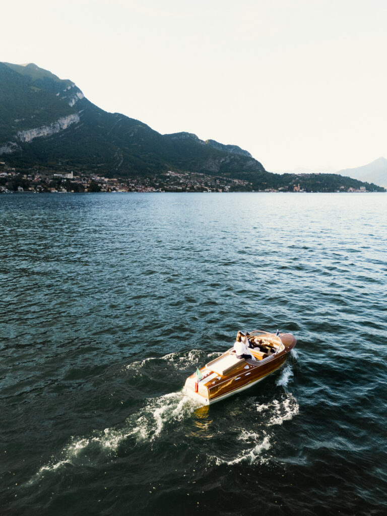 A sweeping aerial-style wide shot looking down at the classic wooden Riva speedboat cutting through the deep blue waters of Lake Como, the bride and groom tiny figures visible on the bow, dwarfed by the vast expanse of the lake and the towering sheer rock face of the Alpine mountains rising dramatically along the shoreline, with a lakeside village nestled at their base in the distance.