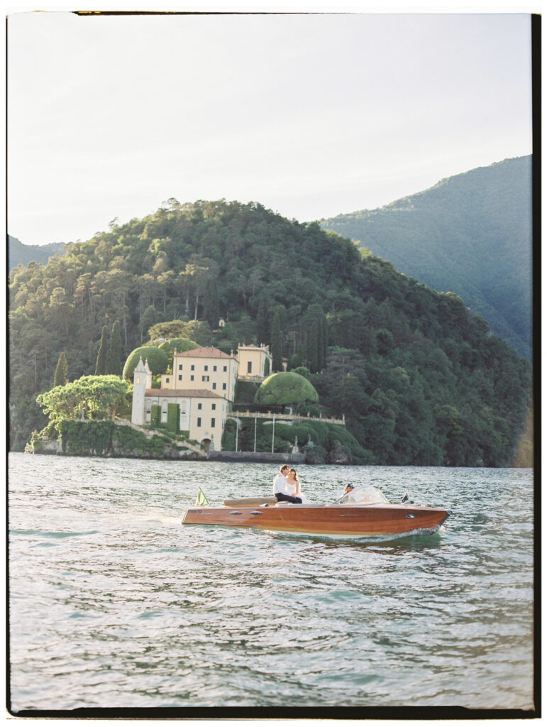 A wide shot of the classic wooden Riva speedboat gliding across the calm waters of Lake Como, tiny against the dramatic backdrop of a grand Italian villa perched on a lush forested peninsula with the steep Alpine mountains rising behind it.