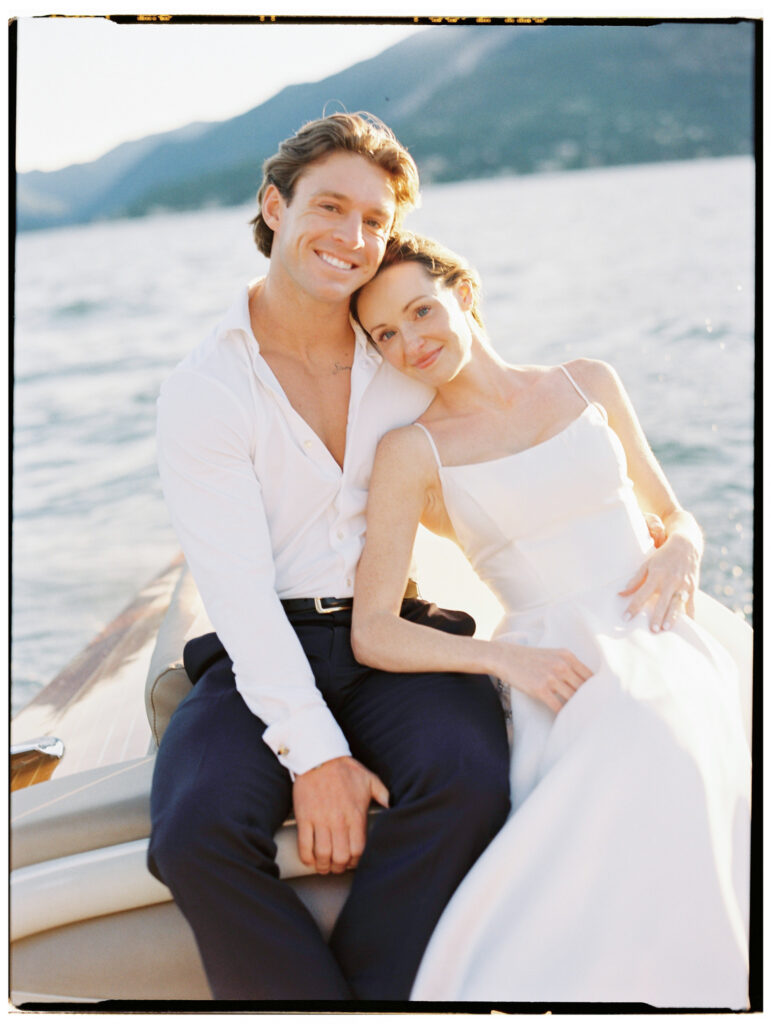The bride rests her head against the groom's chest as they sit together on the Riva speedboat, both smiling warmly and relaxed, with the bright blue waters and lush green hillsides stretching peacefully behind them.