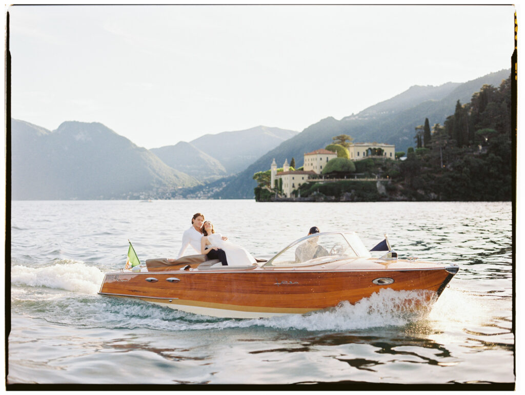 A couple seated closely together on the bow of a classic wooden Riva speedboat gliding across the shimmering waters of Lake Como, the woman leaning back into the man's arms as they look up contentedly. A grand Italian villa nestled among cypress trees and lush hillside greenery rises dramatically behind them, with the misty Alpine mountains stretching across the horizon in soft morning light.