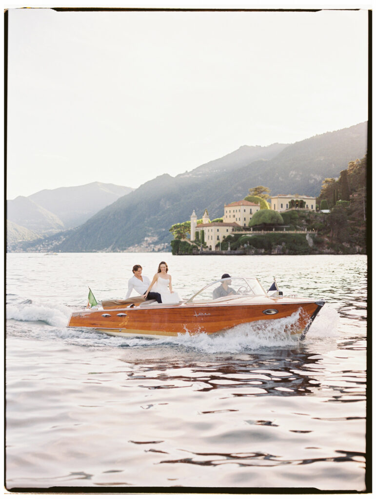 The bride and groom seated together on the bow of a classic wooden Riva speedboat gliding across the sparkling waters of Lake Como at golden hour, with a grand Italian villa nestled among cypress trees and lush green hillsides rising behind them and the Alpine mountains stretching across the horizon.