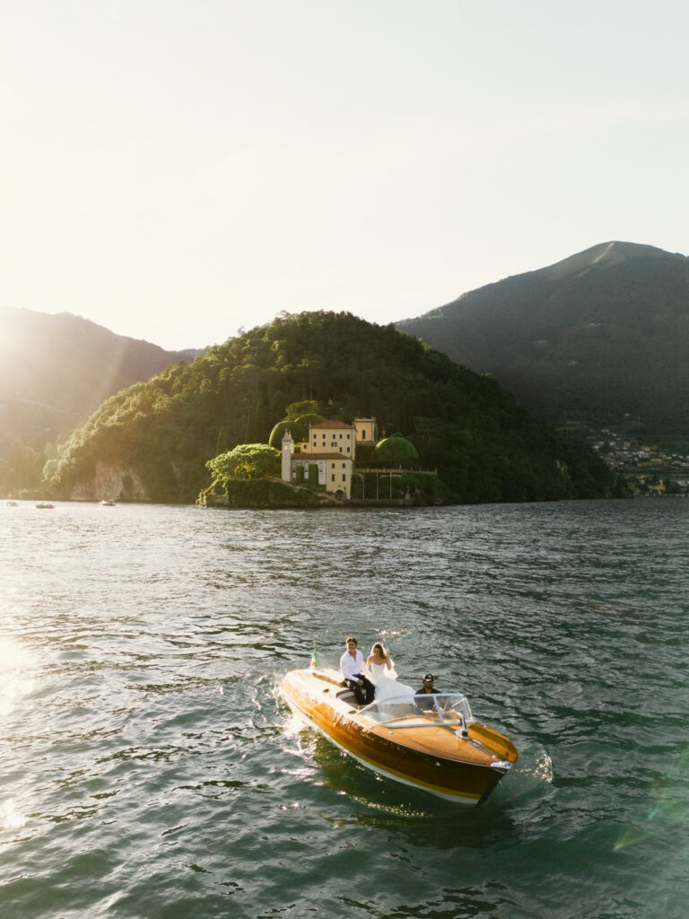 A wide atmospheric shot of the classic wooden Riva speedboat gliding across the golden-lit waters of Lake Como at sunset, with a grand Italian villa dramatically perched on a forested hillside peninsula rising behind it and the Alpine mountains glowing in the warm evening light.