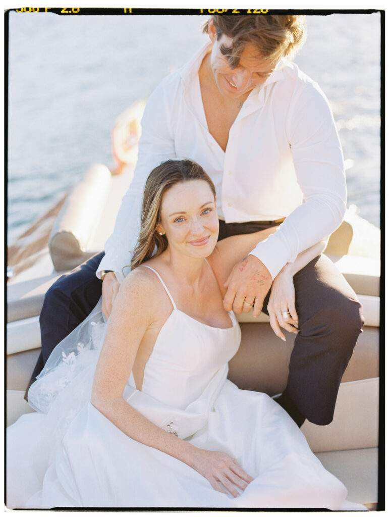 The bride in her white spaghetti-strap wedding gown sits nestled against the groom on the bow of the classic wooden Riva speedboat, smiling softly as he holds her close, with the calm golden waters of Lake Como stretching behind them.