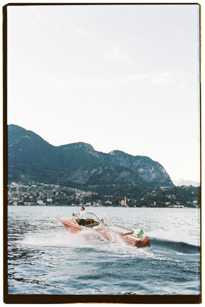 A wide minimalist shot of the classic wooden Riva speedboat, tiny against the dramatic scale of the steep Alpine mountains and a lakeside town visible along the distant shoreline beneath a pale sky.