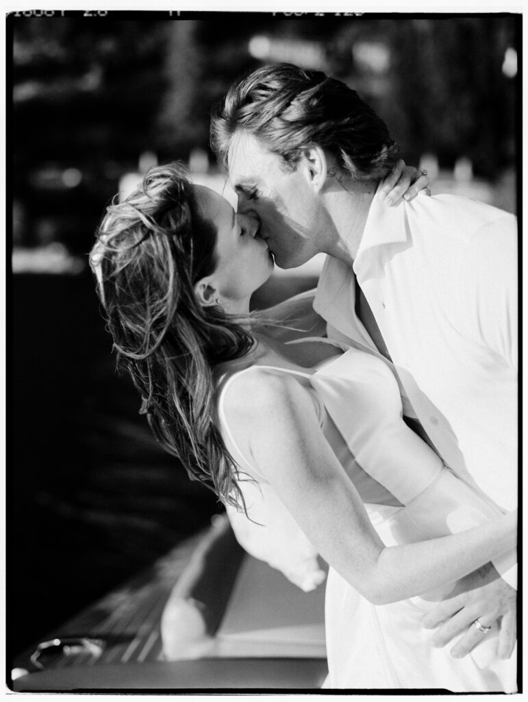A dramatic black-and-white close-up film photograph of the bride and groom leaning in for a kiss aboard the Riva speedboat, their faces nearly touching, hair windswept, captured in a deeply intimate and cinematic moment on Lake Como.