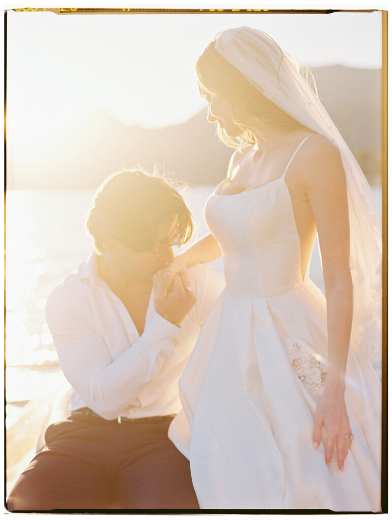 A breathtaking golden hour film photograph of the groom kneeling before the bride on the shores of Lake Como, kissing her hand while she looks down at him tenderly, her white spaghetti-strap gown and cathedral veil glowing in the intense backlit sunlight flooding the scene.