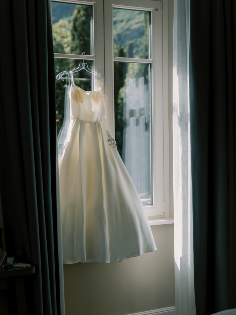 The bride's elegant sleeveless white wedding gown hanging from a hanger against an open window, with the dramatic green mountains and shimmering blue waters of Lake Como framed beautifully in the background through teal curtains.