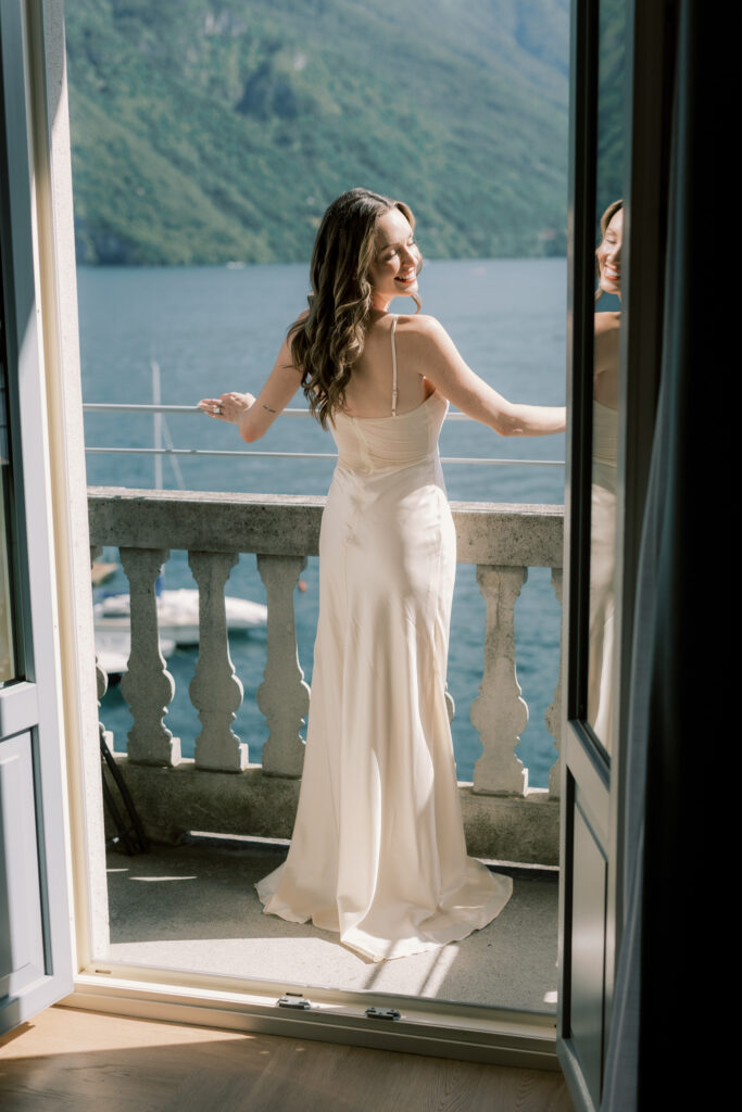 The bride in her sleek white satin wedding gown standing in an open French door balcony, arms outstretched and eyes closed as she takes in the breathtaking view of Lake Como's shimmering blue waters and lush green hillsides behind her.