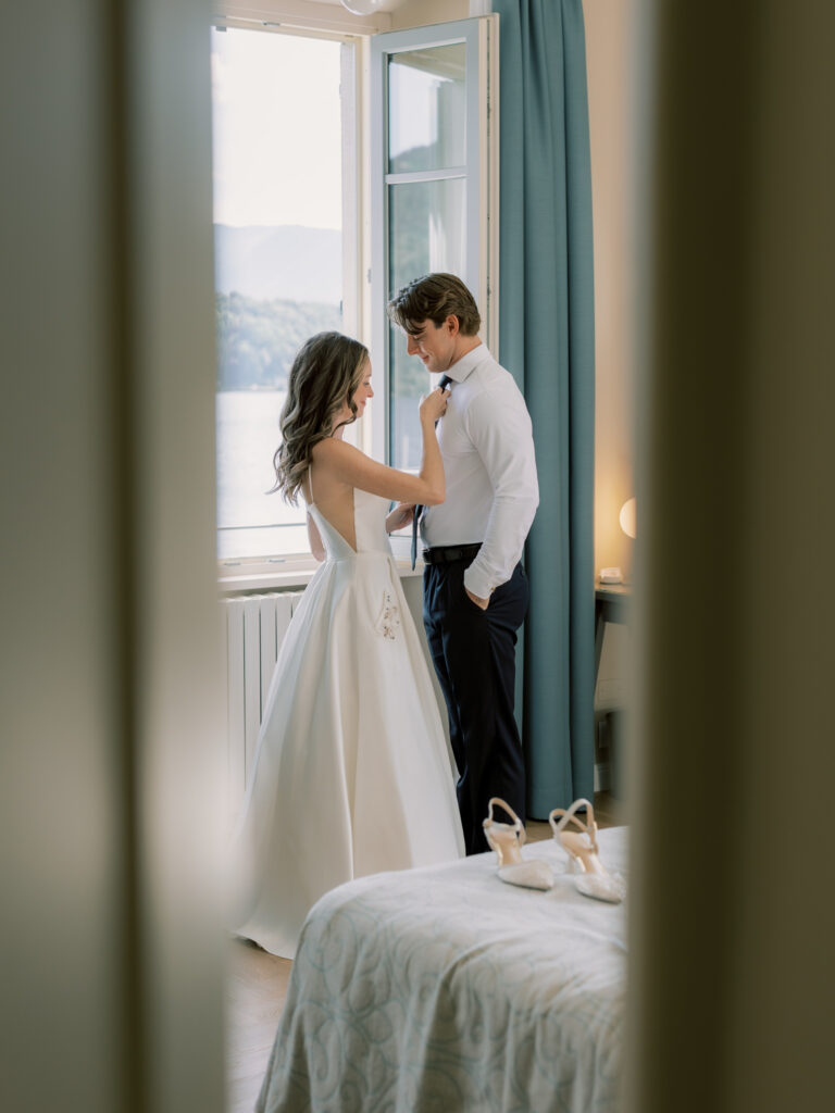 The bride helping the groom knot his tie in a tender, quiet getting-ready moment inside their Lake Como hotel room, both looking down with soft smiles, the bride's flowing white gown and the lake view through the window visible behind them.