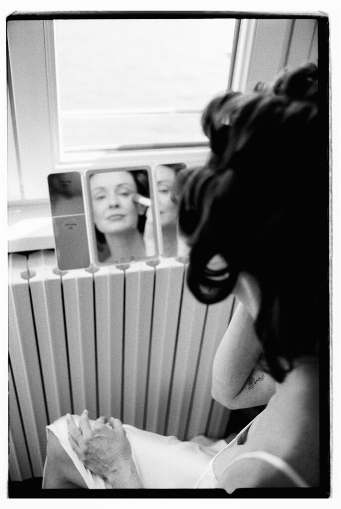 A black-and-white close-up film photograph of the bride applying her makeup using a trifold vanity mirror, her dark hair pinned up as she focuses on her reflection in the bright natural light of their Lake Como hotel room.