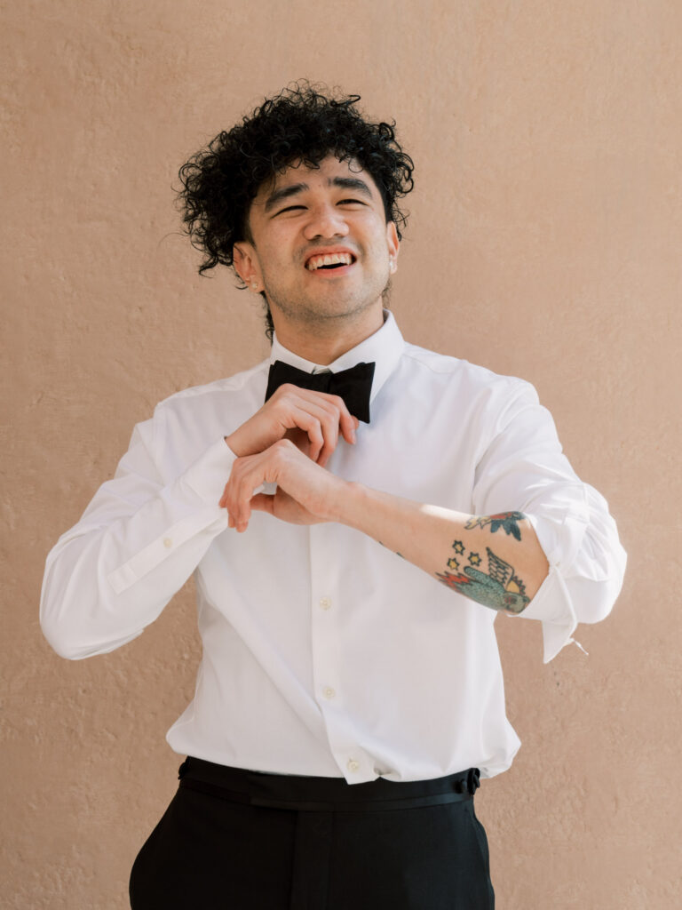 The groom laughs joyfully while adjusting his black bow tie against a warm beige wall, wearing a white dress shirt with his forearm tattoo visible, in a candid and playful getting-ready moment at the One&Only The Palm Dubai wedding.

