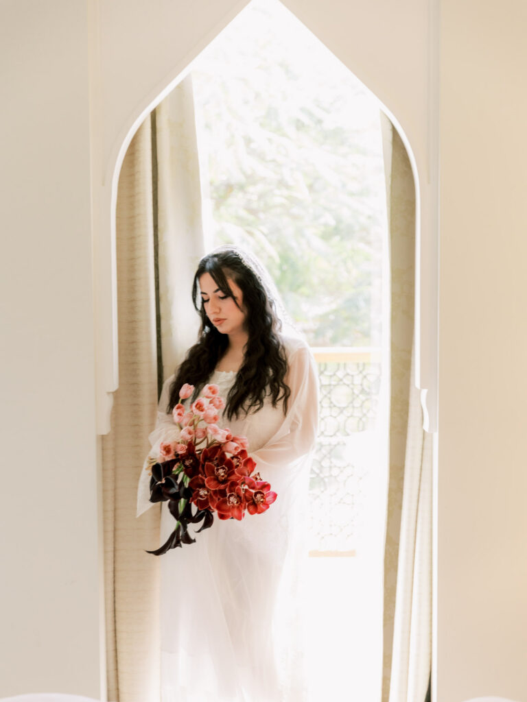The bride in her sheer white bridal robe stands framed within a tall arched white doorway, holding her bouquet of deep pink orchids and black calla lilies, her long dark hair cascading over her shoulders in a dreamy, editorial getting-ready portrait.