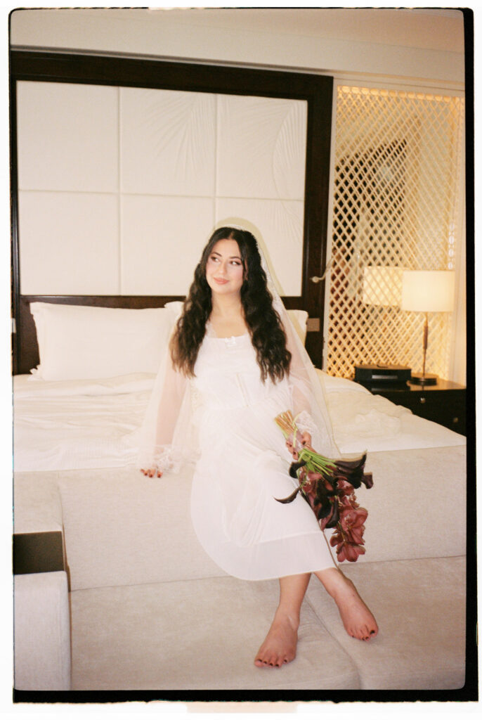 A film photograph of the bride sitting barefoot on the edge of the plush white bed in her One&Only The Palm Dubai hotel suite, wearing her sheer white bridal robe and cathedral veil, holding her deep pink and black bouquet, with the luxurious quilted headboard and decorative lattice wall panel glowing warmly behind her.