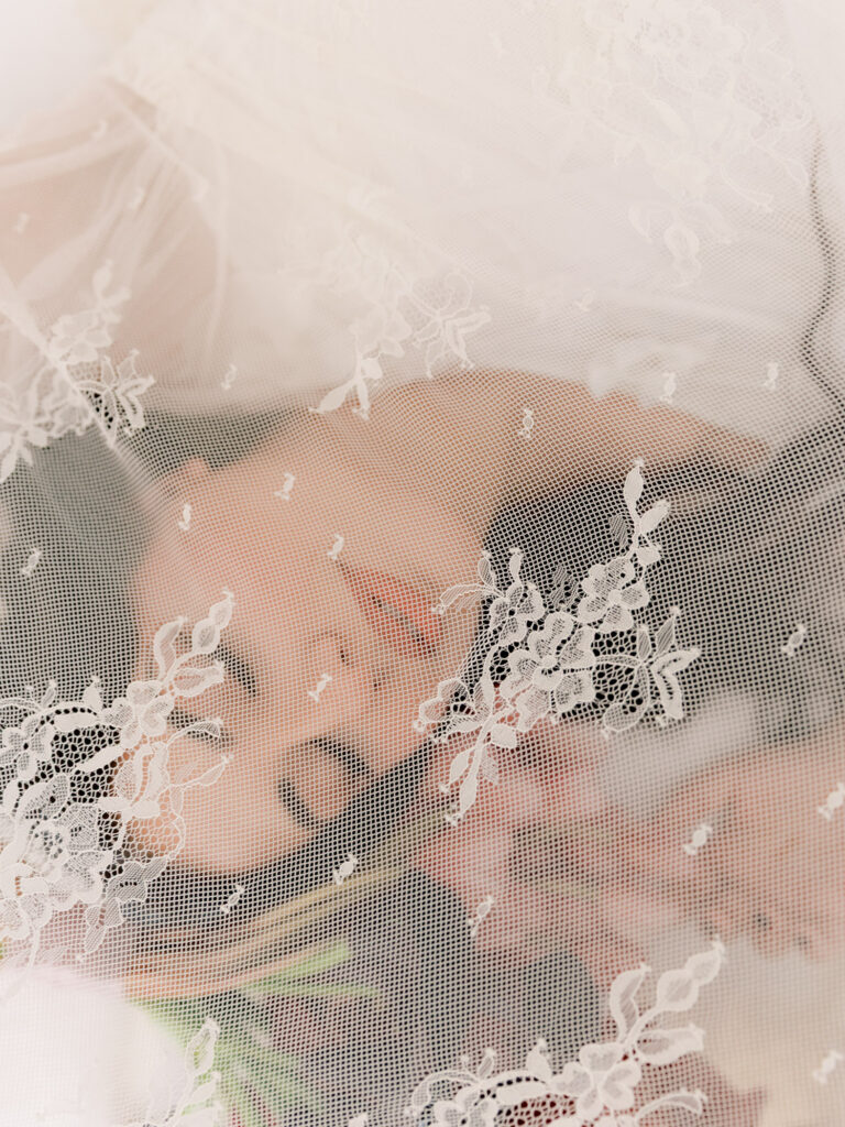A dreamy close-up detail of the bride's face softly visible through her intricate floral lace veil, her pink orchid bouquet blurred in the warm background, creating an ethereal and romantic bridal portrait at One&Only The Palm Dubai wedding.